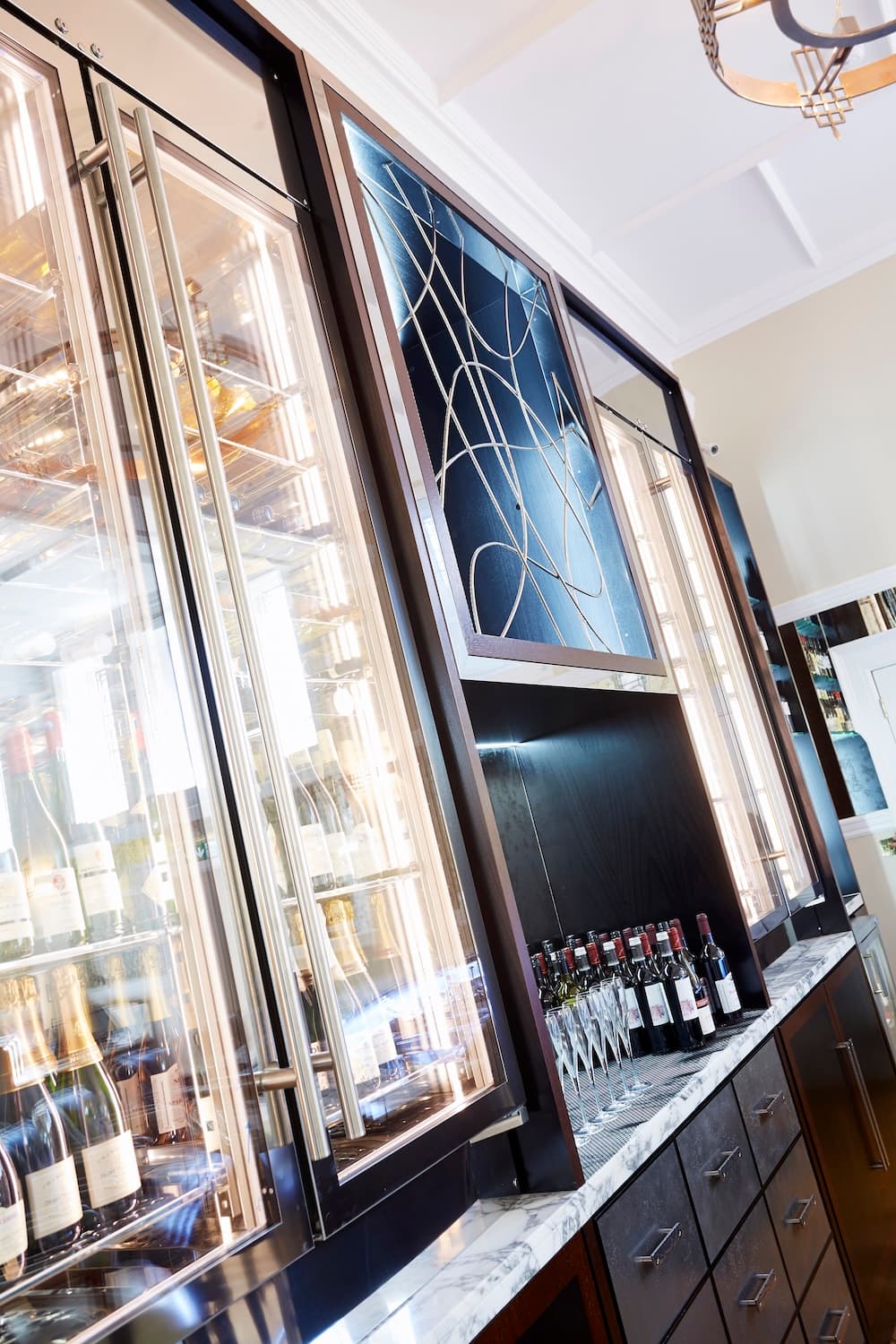 A wide shot of a besopke wooden wine cabinet holding bottles of wine and wine glasses hanging with glass cupboards and a mirror