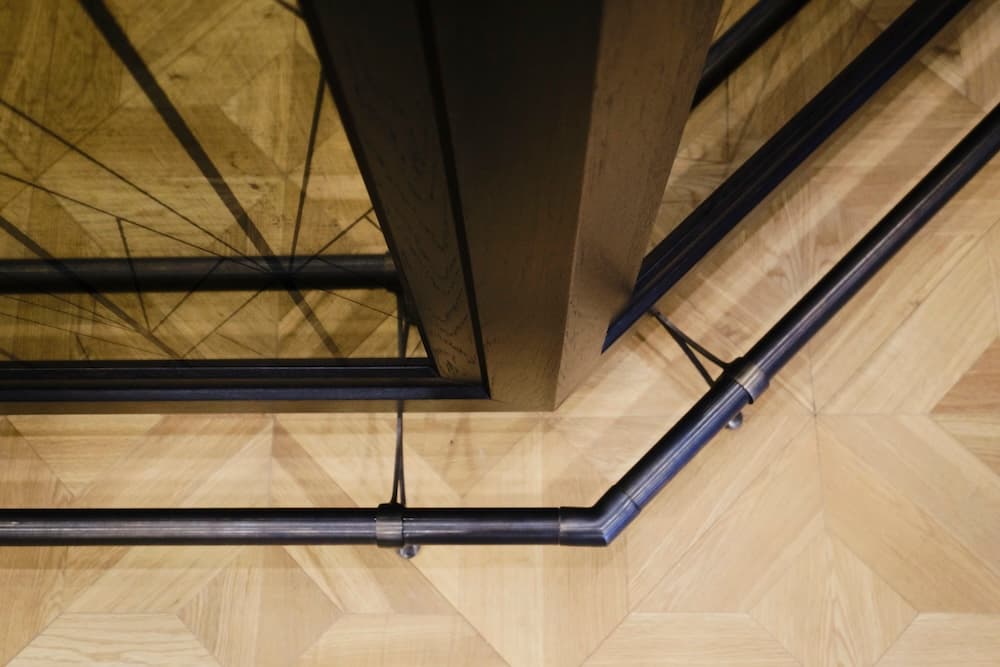 High-angle detail of a precision-engineered dark metal bar footrail mounted against a geometric etched glass panel and light oak parquet flooring.
